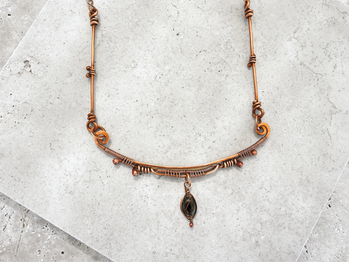 A necklace with a combination of copper wire and beads, ending with a Tiger's Eye pendant drop from a curved copper bar.